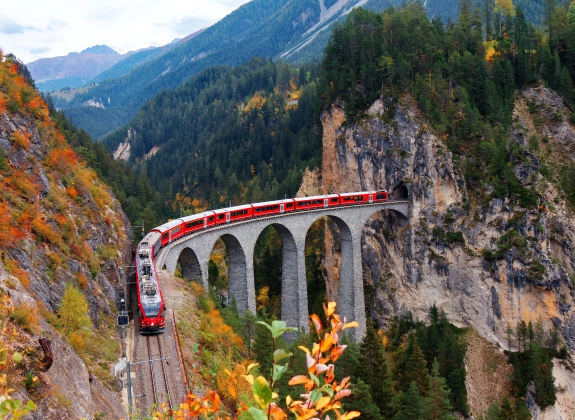 Glacier Express train coming out of the tunnel in a cliff crossing famous Landwasser Viaduct over a deep gorge with fall colors on the rocky mountainside in Filisur, Grisons, Switzerland