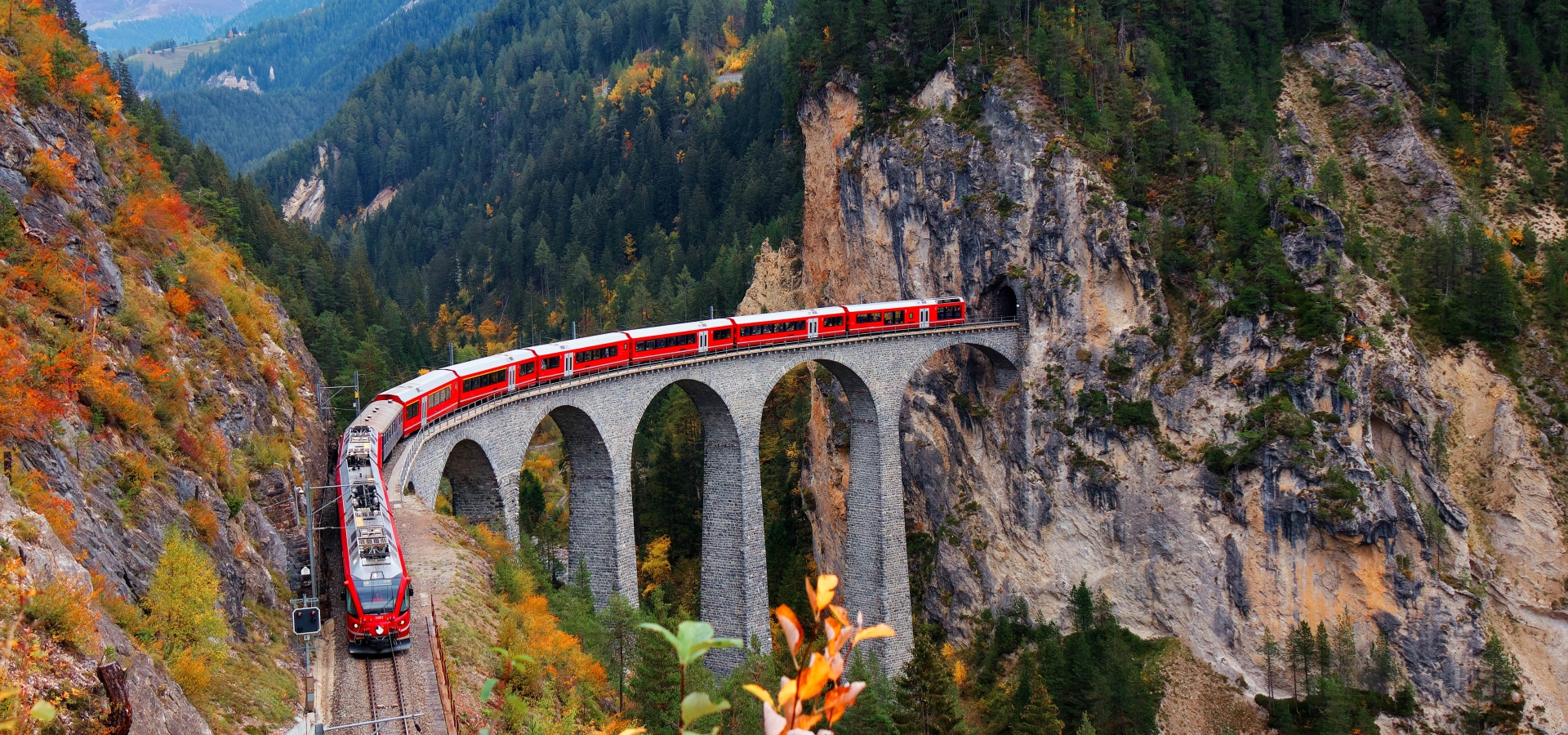 Glacier Express train coming out of the tunnel in a cliff crossing famous Landwasser Viaduct over a deep gorge with fall colors on the rocky mountainside in Filisur, Grisons, Switzerland